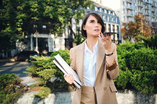 Jeune femme d'affaires séduisante en costume beige avec ordinateur portable enregistrant attentivement un message vocal sur un téléphone portable dans la rue de la ville