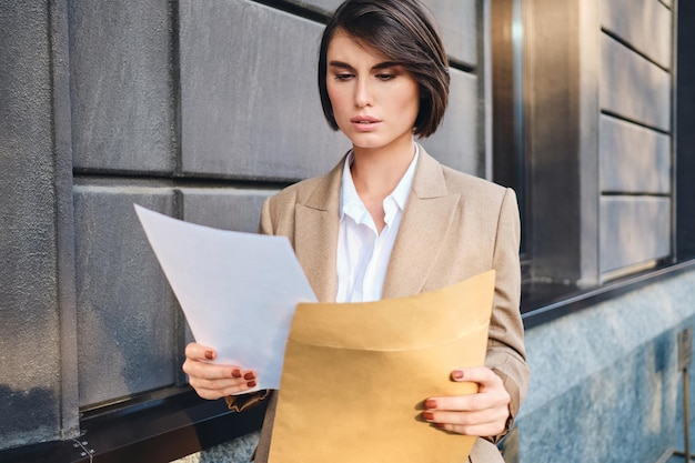 Jeune femme d'affaires sérieuse en costume travaillant de manière réfléchie avec des papiers dans la rue de la ville