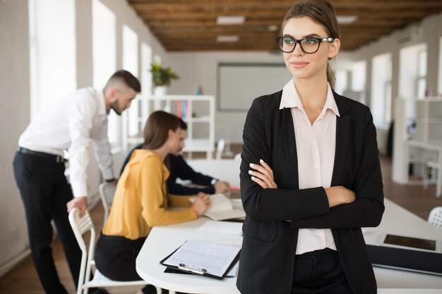 Jeune femme d'affaires souriante en lunettes et chemise regardant rêveusement de côté avec les mains jointes tout en passant du temps au bureau avec des collègues en arrière-plan