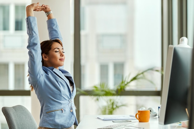 Photo gratuite jeune femme d'affaires souriante qui s'étire les yeux fermés tout en travaillant sur un ordinateur au bureau