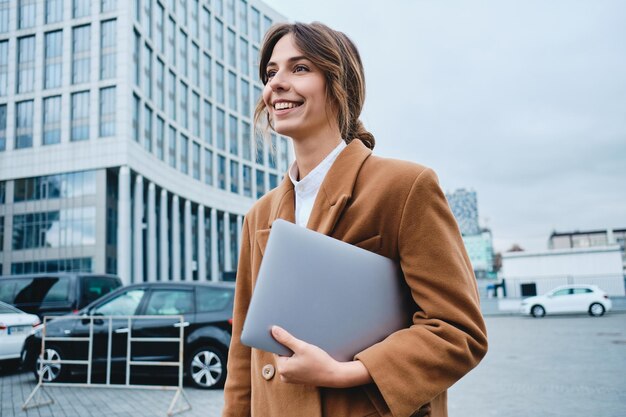 Jeune femme d'affaires souriante séduisante en manteau avec ordinateur portable regardant joyeusement au loin tout en marchant dans la rue de la ville