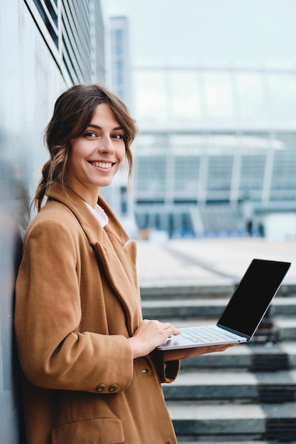 Jeune femme d'affaires souriante séduisante en manteau regardant joyeusement à huis clos travaillant sur un ordinateur portable dans la rue de la ville
