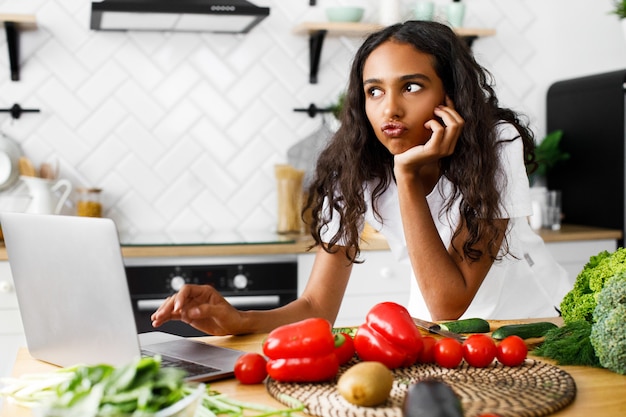 Jeune femme africaine a un air réfléchi en tapant quelque chose dans un ordinateur portable sur un bureau de cuisine avec des légumes
