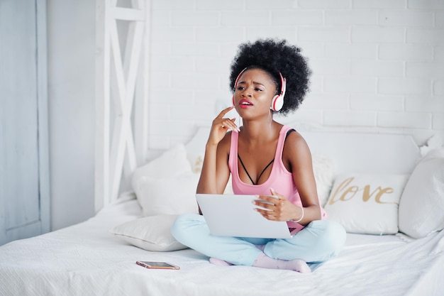 Photo gratuite jeune femme afro-américaine assise dans son lit tout en travaillant sur un ordinateur portable et en écoutant de la musique sur des écouteurs