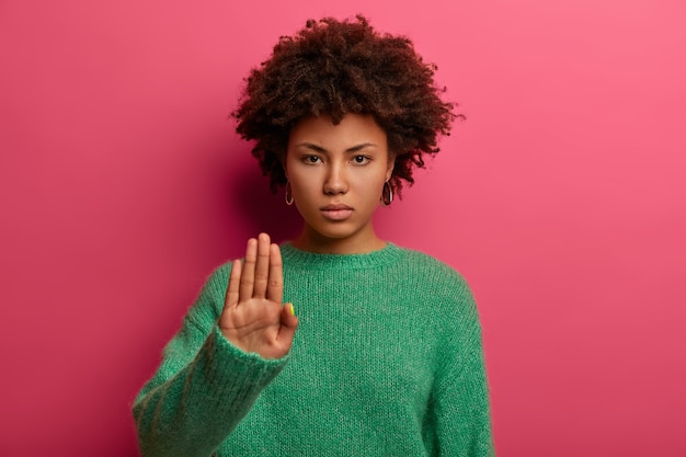 Jeune femme afro-américaine aux cheveux bouclés sérieux fait signe d'arrêt avec paume, porte un pull vert, démontre l'interdiction et la restriction, refuse quelque chose, les modèles contre le mur rose, dit non
