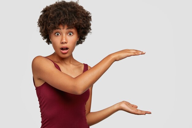 Une jeune femme afro-américaine choquée montre quelque chose de grand avec les mains, ouvre la bouche avec surprise, montre la hauteur de la statue
