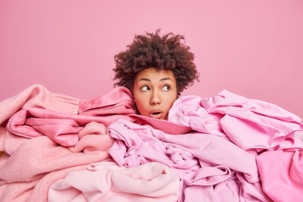 Une jeune femme afro-américaine choquée ne montre que la tête enfouie dans un gros tas de vêtements détourne le regard avec une expression abasourdie recueille des vêtements pour un don isolé sur du rose