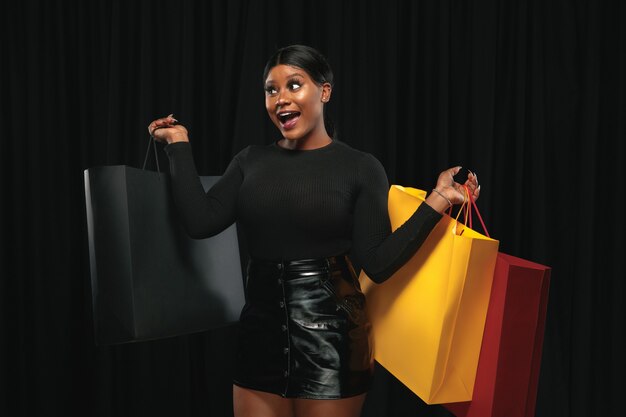 Jeune femme afro-américaine faisant du shopping avec des packs colorés sur un mur noir.