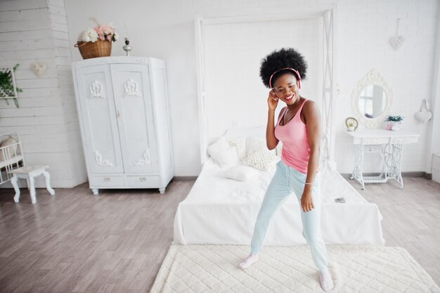 Jeune femme afro-américaine en maillot rose dansant et écoutant de la musique sur des écouteurs dans sa chambre blanche