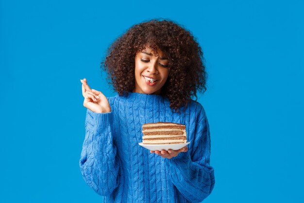 Jeune femme afro-américaine mignonne et idiote d'espoir en pull, avec coupe de cheveux afro bouclée, croiser les doigts bonne chance, prier et tenant une assiette avec un délicieux gâteau aux gros morceaux, mur bleu debout.