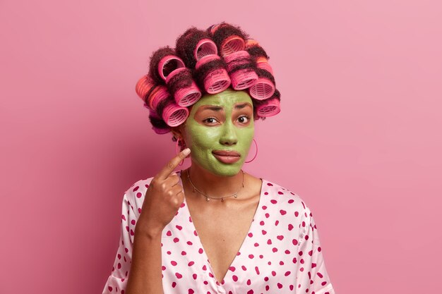 Jeune femme afro-américaine montre comment elle se soucie de la peau, applique un masque vert, attend l'effet de rajeunissement, applique des rouleaux de cheveux, se prépare pour la fête, porte une robe décontractée, pose à l'intérieur