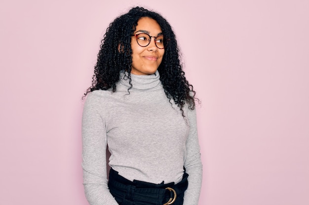 Photo gratuite jeune femme afro-américaine portant un pull à col roulé et des lunettes sur fond rose souriant regardant sur le côté et regardant loin en pensant