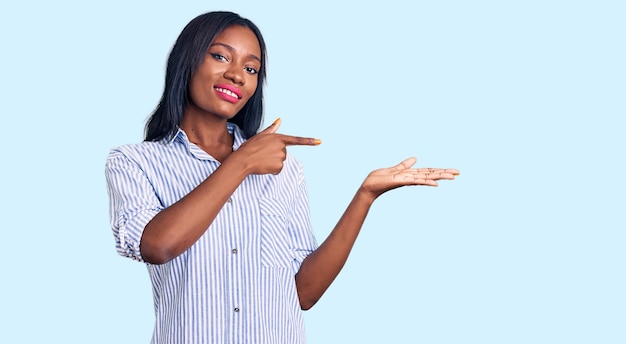 Jeune femme afro-américaine portant des vêtements décontractés étonnée et souriante à la caméra tout en se présentant avec la main et en pointant du doigt