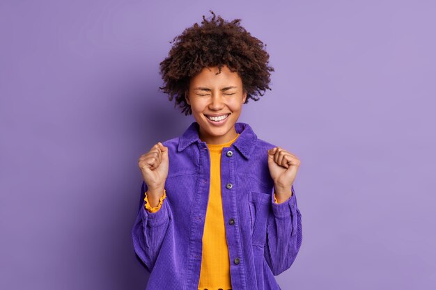 Jeune femme afro-américaine ravie positive lève les poings fermés célèbre quelque chose avec triomphe vêtu d'une veste à la mode