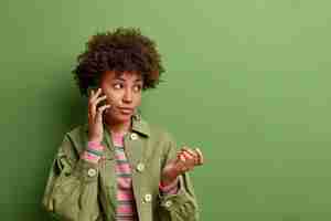 Photo gratuite une jeune femme afro-américaine réfléchie lève la main a une conversation téléphonique considère les informations entendues par l'interlocuteur porte des vêtements élégants isolés sur un mur vert vif