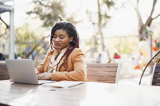 Jeune femme afro-américaine souriante assise à la table dans un café de la rue et utilisant un ordinateur portable