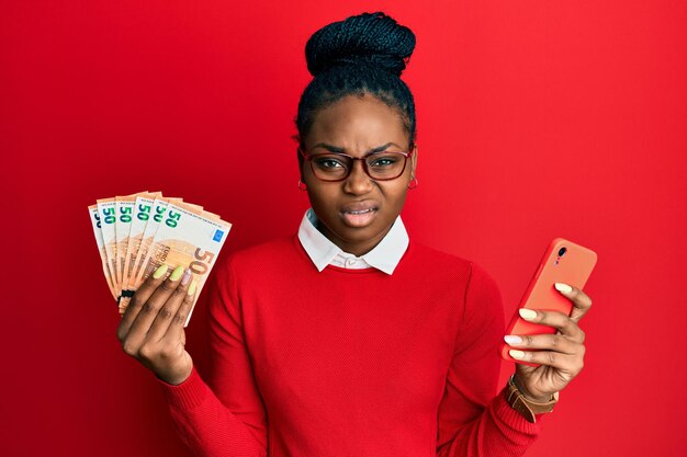 Jeune femme afro-américaine utilisant un smartphone tenant des billets en euros expression désemparée et confuse. notion de doute.