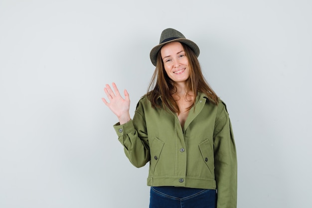 Jeune femme agitant la main pour dire au revoir en veste, pantalon, chapeau et à la joyeuse. vue de face.