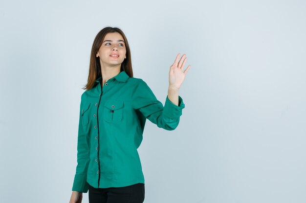 Jeune femme agitant la main pour saluer tout en regardant en arrière en chemise verte et à la joyeuse, vue de face.