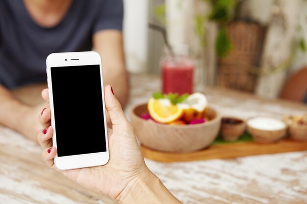 Jeune femme à l'aide de téléphone intelligent tout en déjeunant au café. Caucasienne femme tenant un appareil électronique avec écran blanc avec espace de copie pour votre contenu promotionnel.