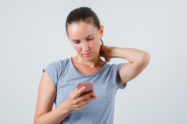 Photo gratuite jeune femme à l'aide de téléphone portable avec la main sur le cou en t-shirt gris, vue de face.