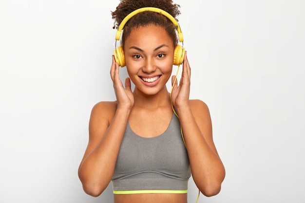 Jeune femme aime la musique comme motivation personnelle, garde les deux mains sur les écouteurs, sourit agréablement, porte un soutien-gorge de sport gris