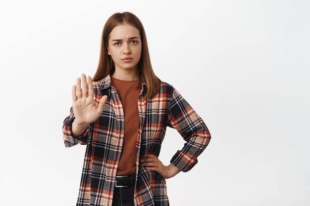 La jeune femme a l'air épuisée et lève la main en bloc, dit non, refuse d'interdire ou rejette quelque chose de décevant, disant de rester à l'écart, debout contre un mur blanc.