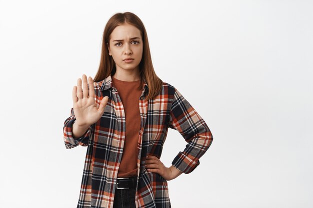 La jeune femme a l'air épuisée et lève la main en bloc, dit non, refuse d'interdire ou rejette quelque chose de décevant, disant de rester à l'écart, debout contre un mur blanc.