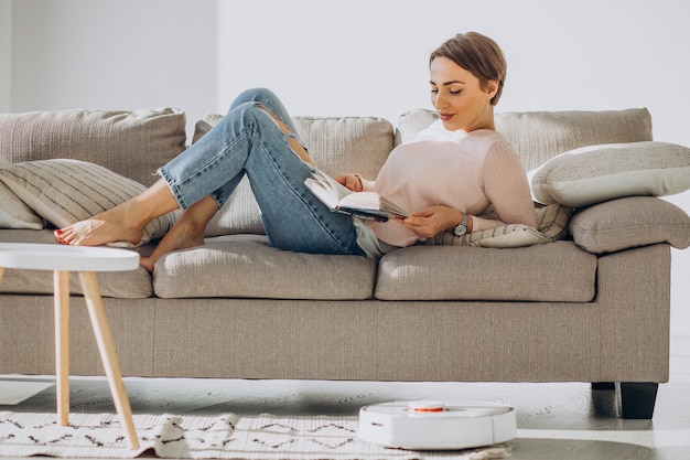 Jeune femme allongée sur un canapé et lisant un livre pendant que l'aspirateur robot fait le ménage