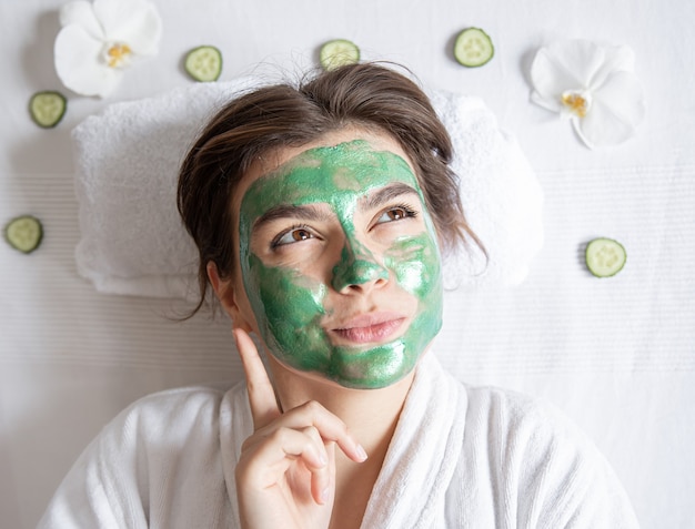 Une jeune femme amusante avec un masque cosmétique vert sur le visage se repose en position allongée dans le salon de spa, vue de dessus.