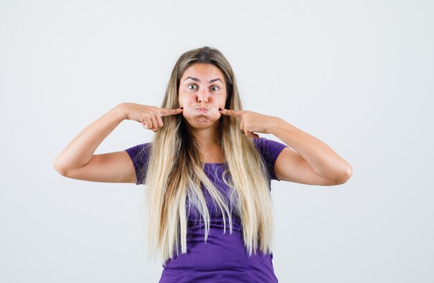 Jeune femme en appuyant sur les doigts sur les joues soufflées en t-shirt violet, vue de face.