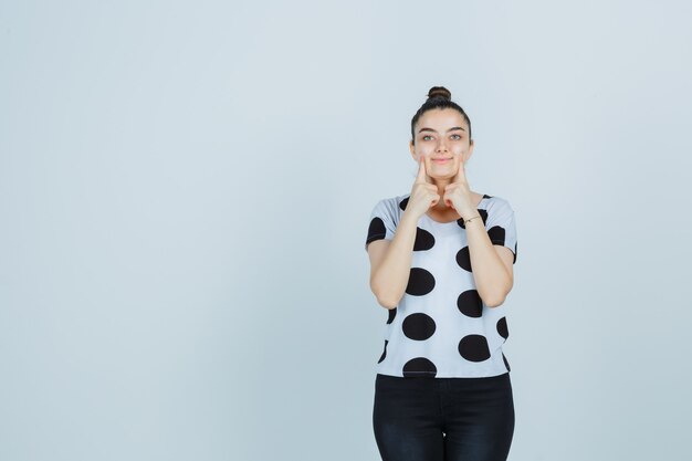 Jeune femme en appuyant sur les doigts sur les joues en t-shirt, jeans et à la jolie vue de face.