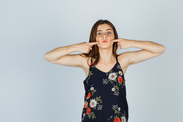 Jeune femme en appuyant sur les joues avec les doigts en blouse et à la perplexité. vue de face.