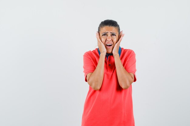 Jeune femme en appuyant sur les oreilles avec les mains en t-shirt rouge et à l'ennui. vue de face.