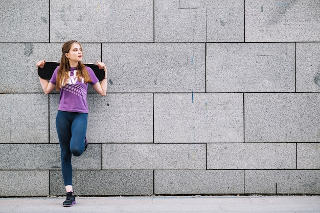 Jeune femme appuyée contre un mur urbain carrelé gris debout sur une jambe