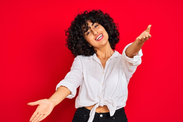 Photo gratuite jeune femme arabe aux cheveux bouclés portant une chemise décontractée blanche sur fond rouge isolé regardant la caméra en souriant à bras ouverts pour un câlin expression joyeuse embrassant le bonheur