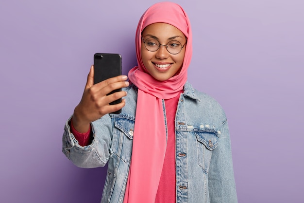 Jeune femme arabe avec une expression joyeuse, porte des lunettes rondes, fait un appel vidéo via cellulaire