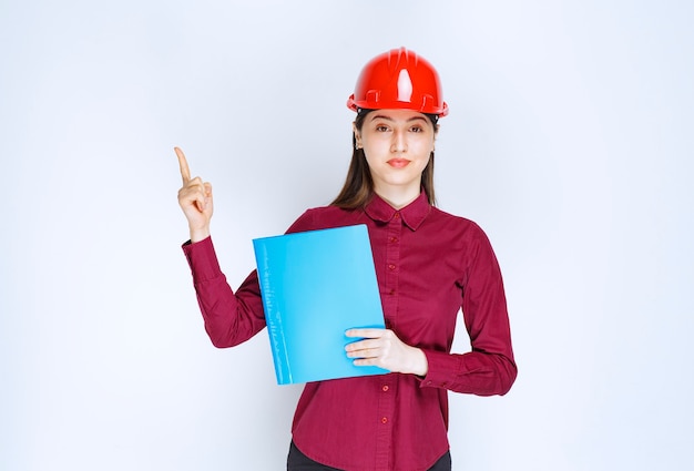 Jeune femme architecte en casque rouge tenant des documents et pointant sur fond blanc.