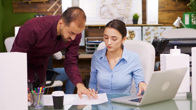 Jeune femme architecte travaillant sur un ordinateur portable et parlant avec le constructeur en chef sur des imprimés bleus.