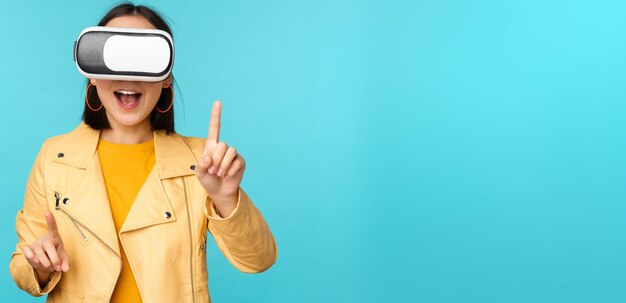 Jeune femme asiatique à l'aide de lunettes de réalité virtuelle à l'aide d'un casque VR debout amusé sur fond bleu Espace de copie