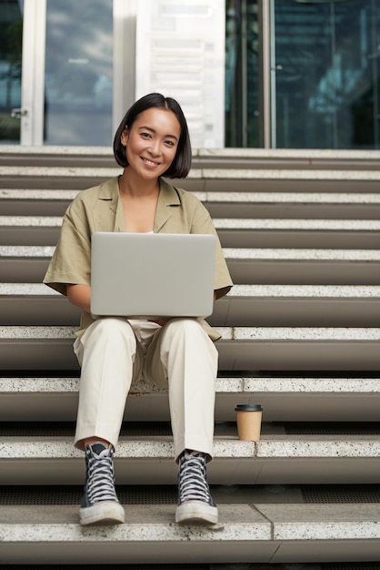 Jeune femme asiatique à l'aide d'un ordinateur portable à l'extérieur jolie fille étudiante avec ordinateur fait ses types de devoirs assis