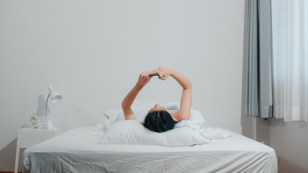 Jeune femme asiatique à l'aide de smartphone, vérification des médias sociaux, sentiment heureux de sourire en position couchée sur le lit après le réveil du matin, belle demoiselle hispanique attrayante souriante se détendre dans la chambre à la maison.