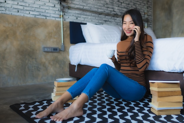 Jeune femme asiatique à l'aide de téléphone mobile intelligent avec livre de lecture et tasse de café dans la chambre