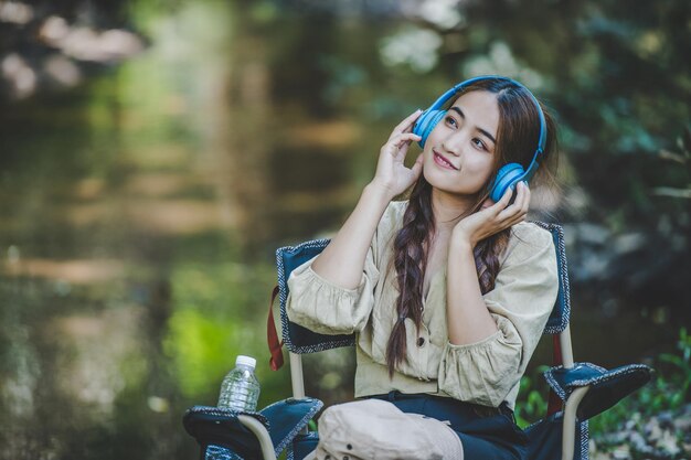 Jeune femme asiatique assise sur une chaise près du ruisseau, écoutant de la musique sur des écouteurs sans fil et utilisant une tablette avec bonheur tout en campant dans l'espace de copie des bois