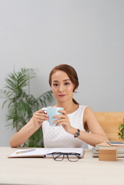 Jeune femme asiatique assise à table à l'intérieur et tenant la tasse