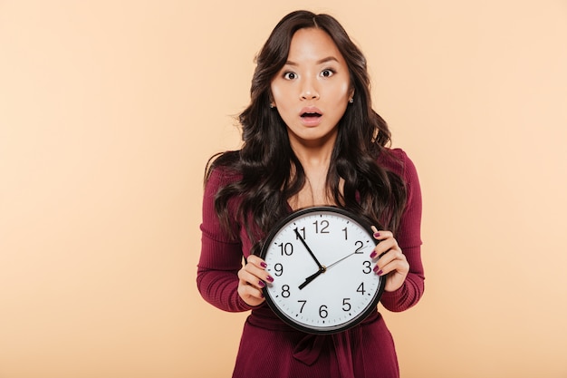 Jeune femme asiatique aux cheveux longs bouclés tenant une horloge montrant près de 8 étant en retard ou manquant quelque chose sur fond de pêche