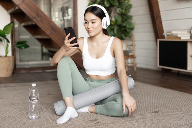 Jeune femme asiatique avec une bouteille d'eau écoutant de la musique ou un podcast dans les écouteurs pendant la remise en forme à la maison ...