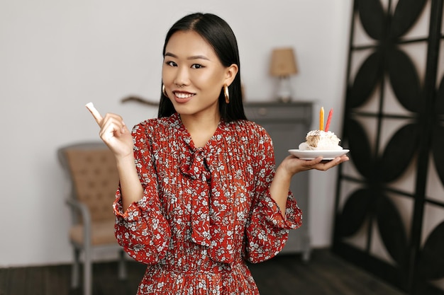 Jeune femme asiatique brune en robe à fleurs tient un délicieux gâteau d'anniversaire crémeux avec des bougies Jolie dame en tenue rouge sourit dans une chambre confortable