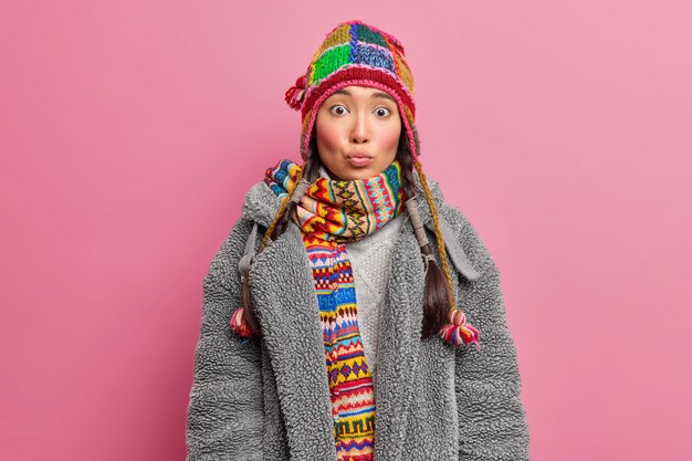 Jeune femme asiatique garde les lèvres arrondies a surpris l'expression porte un bonnet tricoté et une écharpe vêtue d'un manteau de fourrure gris isolé sur un mur rose