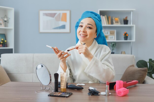 Jeune femme asiatique heureuse souriante avec une serviette sur la tête assise à la coiffeuse à l'intérieur de la maison appliquant des fards à paupières avec miroir faisant la routine de maquillage du matin